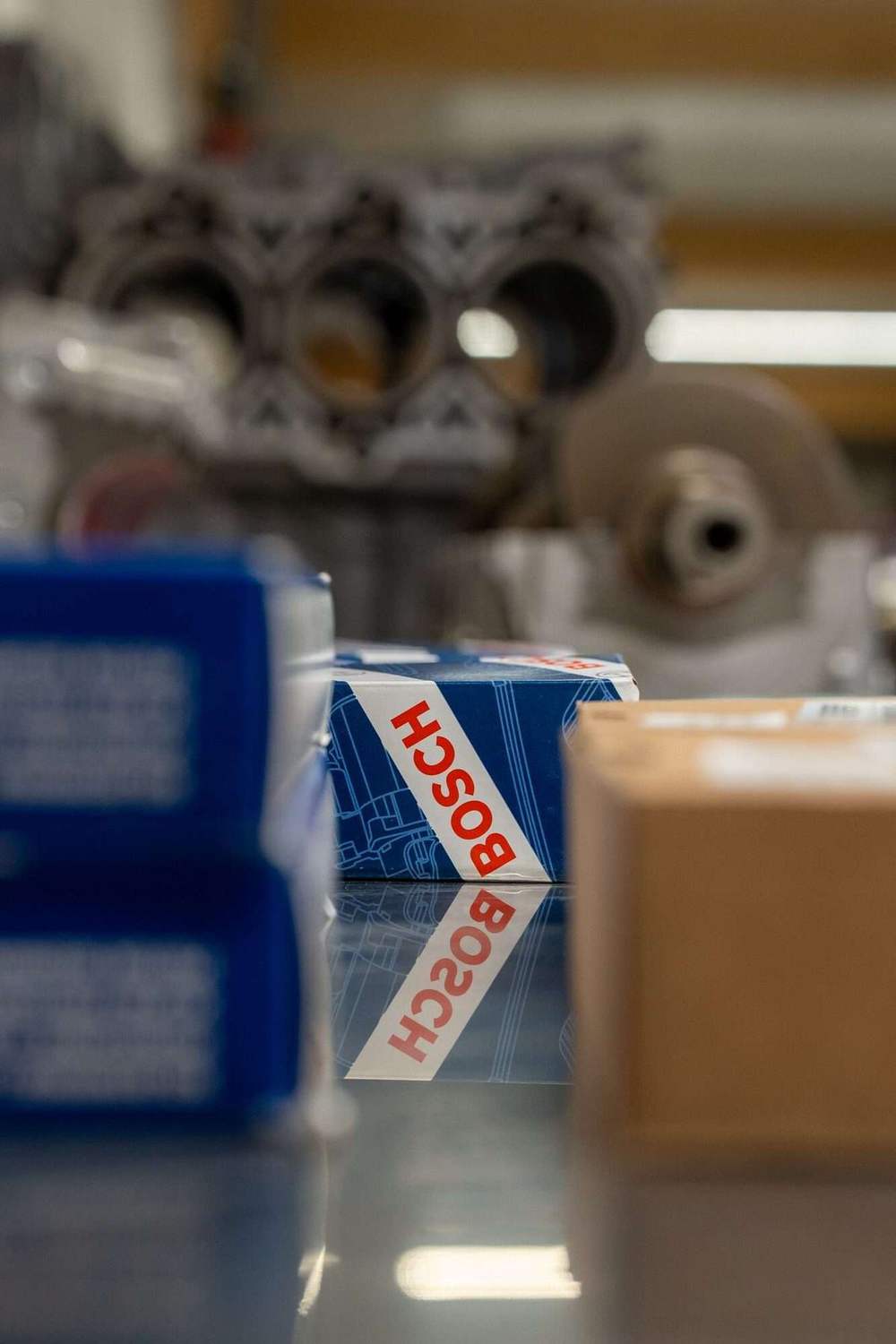 Bosch-Originalteil in Verpackung vor unscharfem Porsche-Motorblock in der Werkstatt von Paintmayer