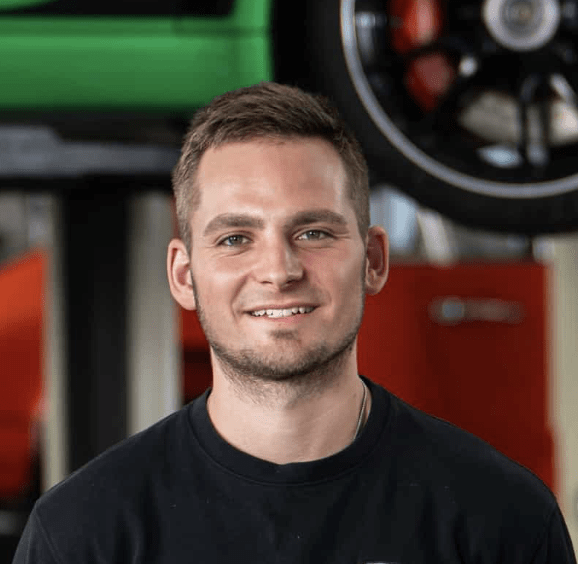 Ein lächelnder Mann mit kurzen braunen Haaren und gestutztem Bart trägt ein schwarzes Hemd und steht in einem Innenraum vor einer Autoausrüstung und einem angehobenen Fahrzeugreifen im Hintergrund.