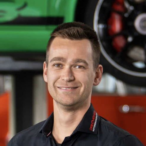 Ein Mann mit kurzen braunen Haaren, der ein schwarzes Hemd mit rotem Kragen trägt, steht in einem Innenraum vor einem grünen Auto und einem großen Rad. Er lächelt und blickt leicht zur Seite.