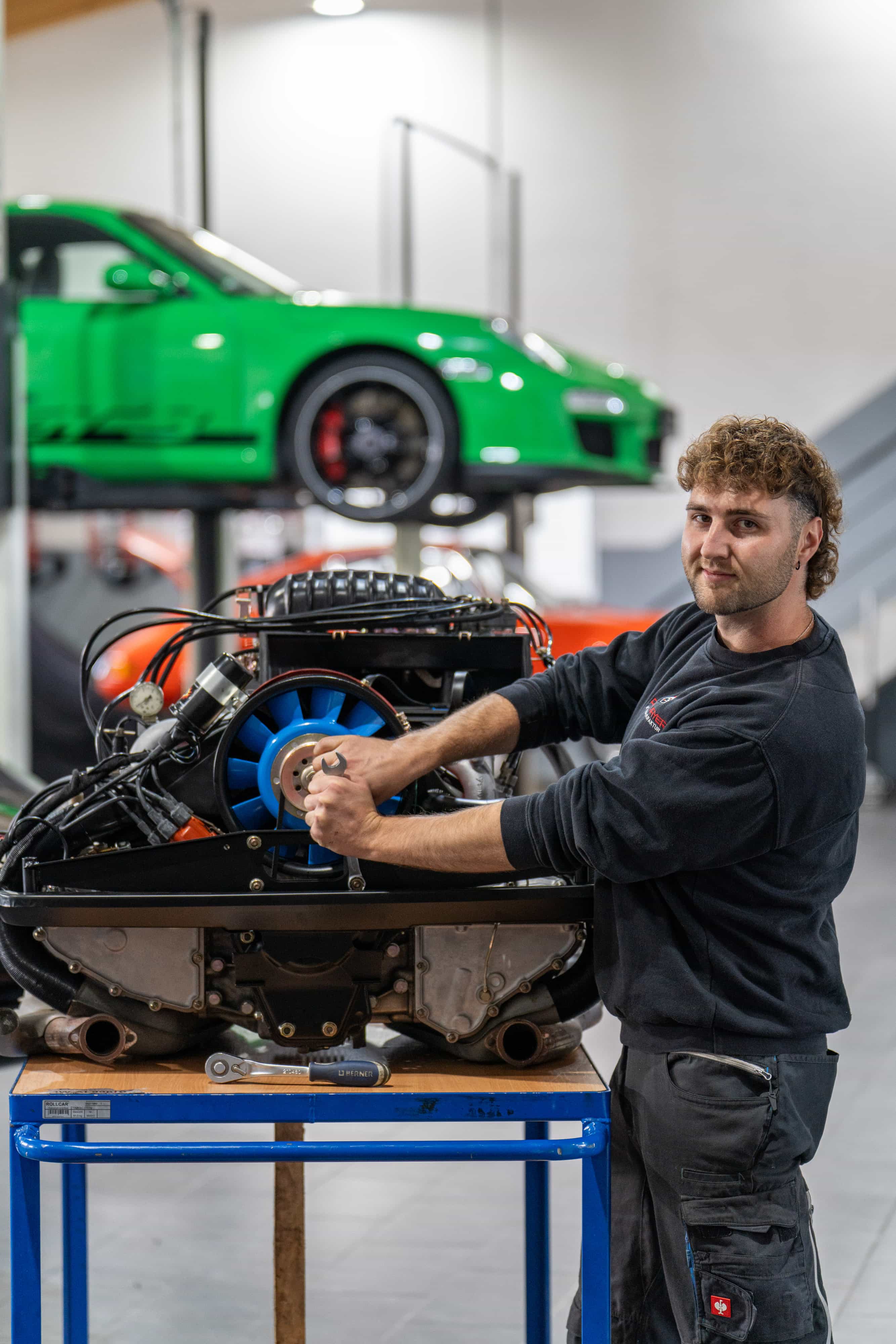 Drei Automechaniker arbeiten in einer hellen Garage an Luxusautos: Einer gießt Flüssigkeit in den Motor eines schwarzen Autos, ein anderer reinigt Motorteile, und ein dritter inspiziert die Unterseite eines gelifteten grünen Sportwagens.