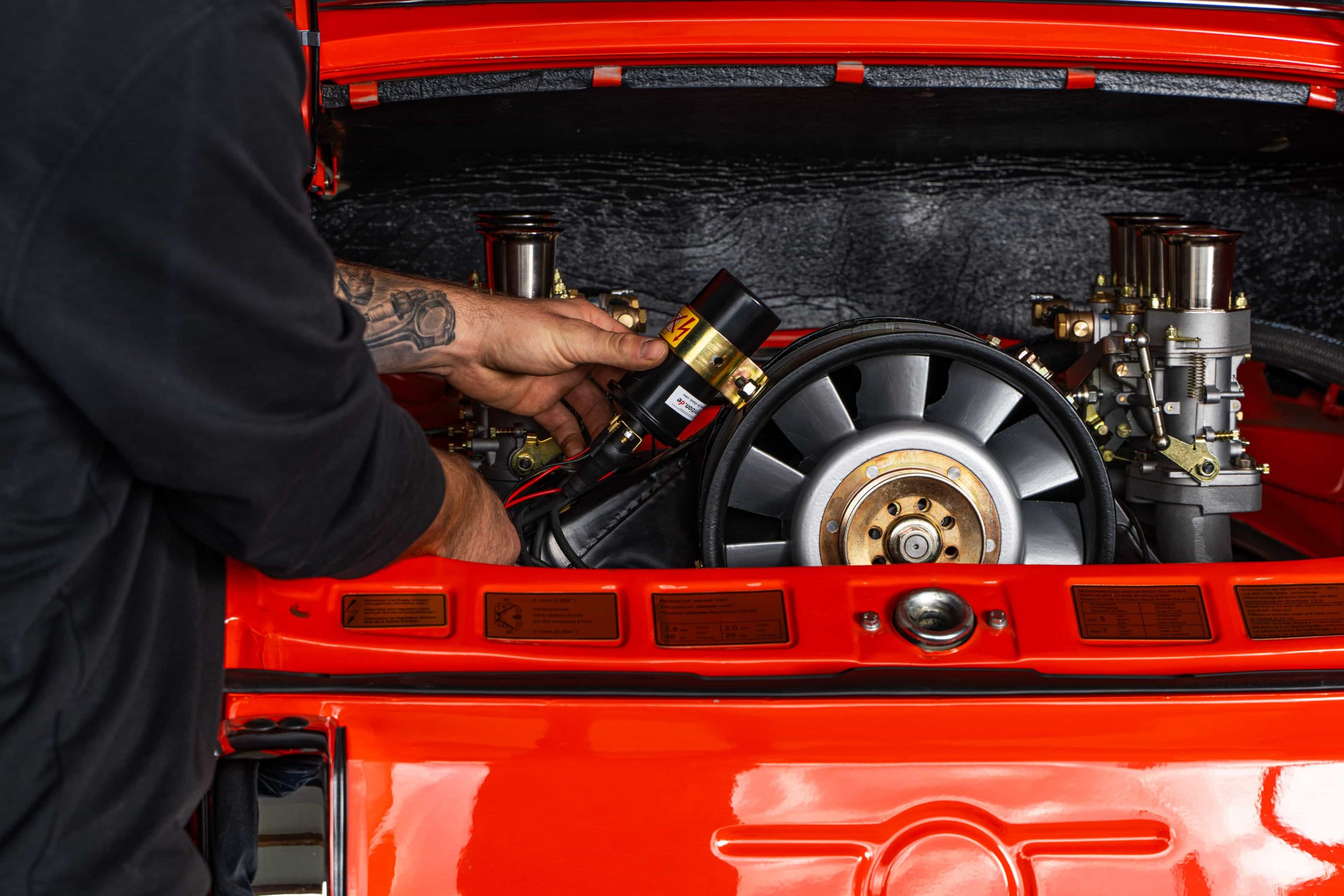 Eine Person mit tätowierten Armen arbeitet am freiliegenden Motor eines roten Oldtimers und stellt ein Bauteil im offenen Kofferraum ein oder baut es ein.
