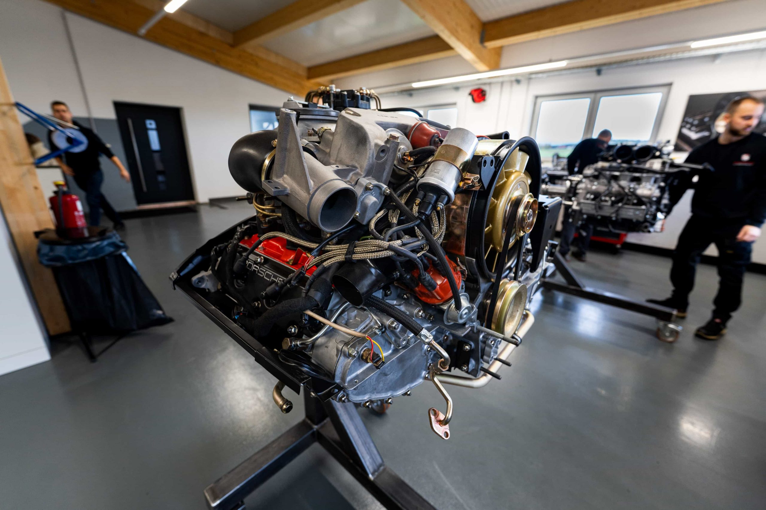 Ein detaillierter, auf einem Ständer montierter Automotor ist in einer Werkstatt ausgestellt, wobei zwei Personen im Hintergrund verschwommen sind. Der Raum hat graue Böden, weiße Wände und sichtbare Holzbalken an der Decke.