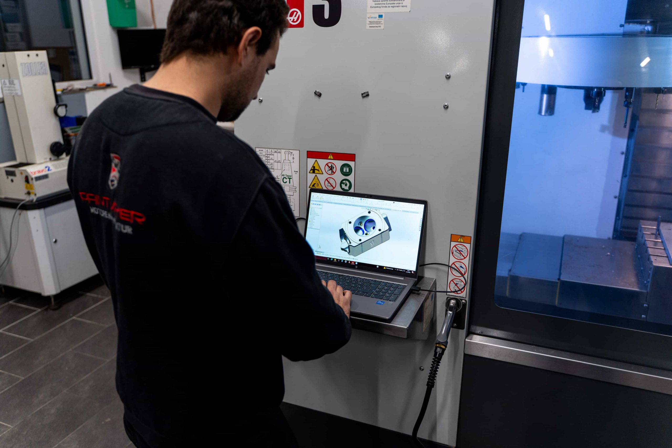 Ein Mann arbeitet mit einem Laptop, auf dem ein 3D-Modell angezeigt wird, neben einer Industriemaschine in einer modernen Werkstatt. Sicherheits- und Warnschilder sind an der Maschine neben ihm zu sehen.