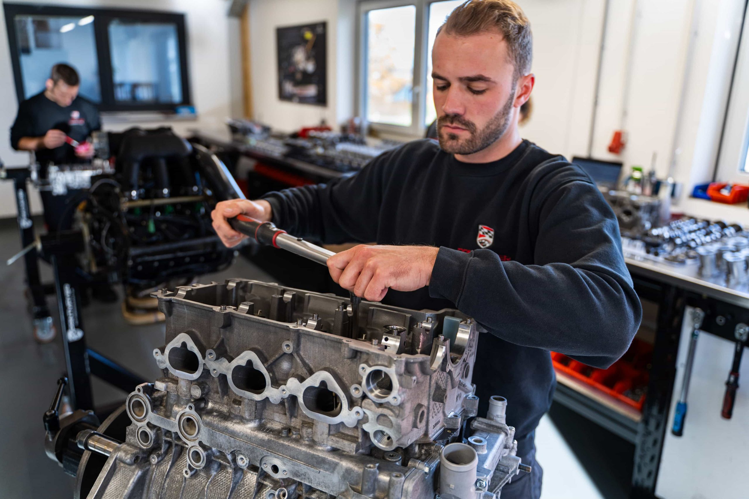 Ein Mann arbeitet mit einem Drehmomentschlüssel an einem Motorblock in einer Werkstatt, während eine andere Person im Hintergrund arbeitet, umgeben von Autoteilen und Werkzeugen.