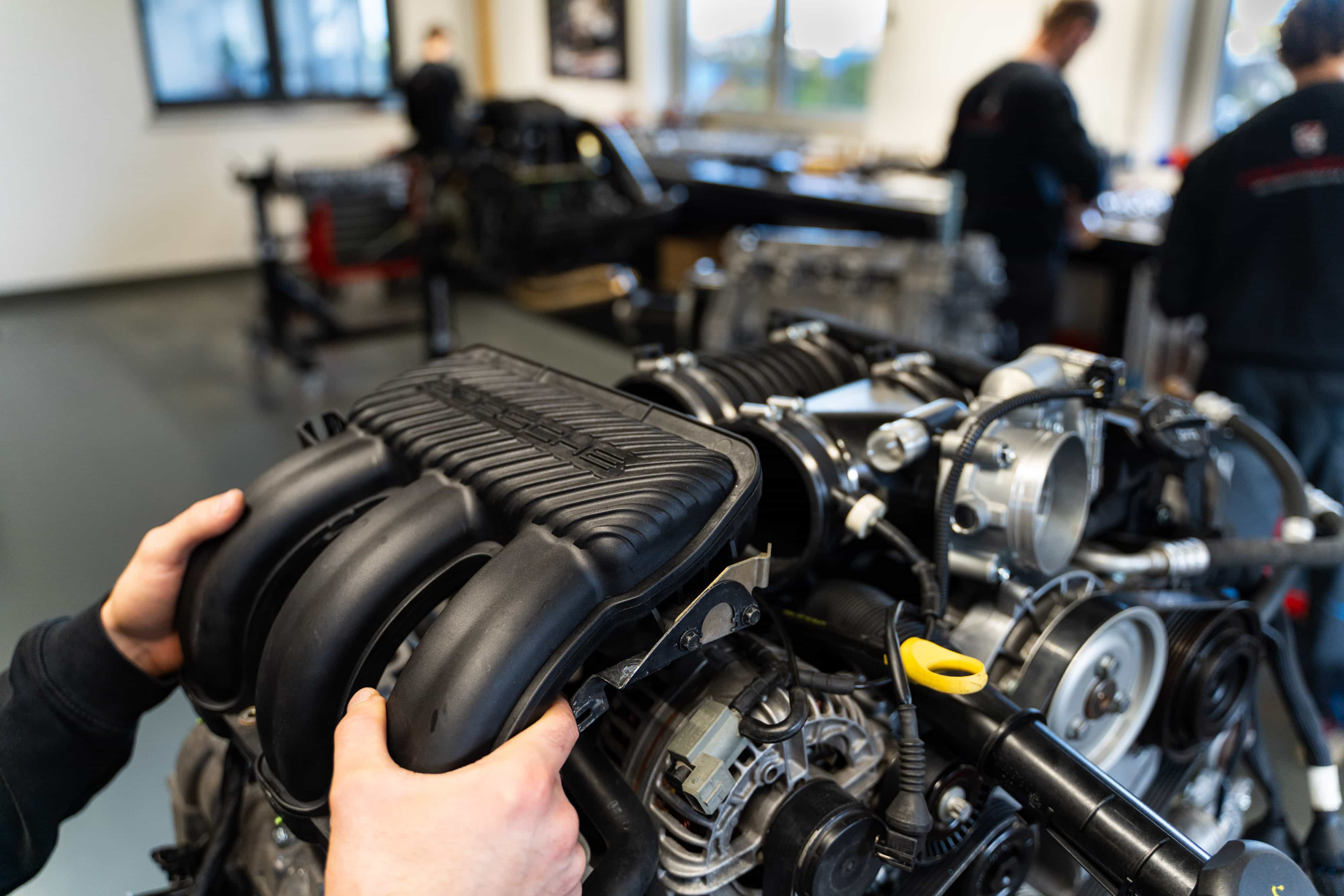 Ein Mann arbeitet mit einem Drehmomentschlüssel an einem Motorblock in einer Werkstatt, während eine andere Person im Hintergrund arbeitet, umgeben von Autoteilen und Werkzeugen.