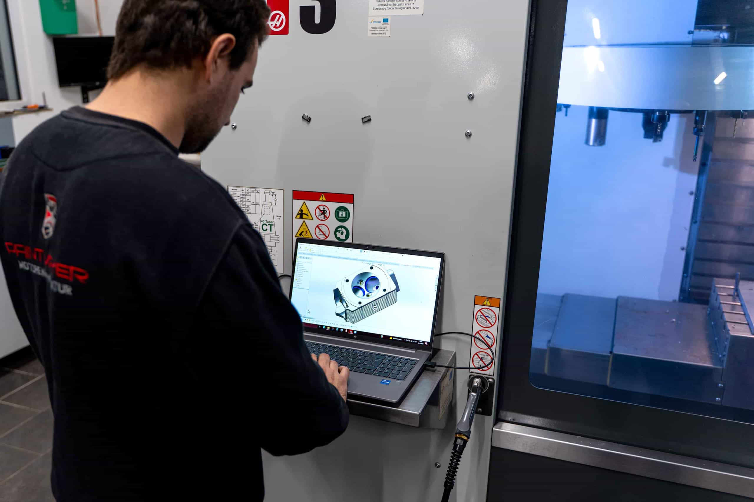 Ein Mann arbeitet mit einem Laptop, auf dem ein 3D-Modell angezeigt wird, neben einer Industriemaschine in einer modernen Werkstatt. Sicherheits- und Warnschilder sind an der Maschine neben ihm zu sehen.