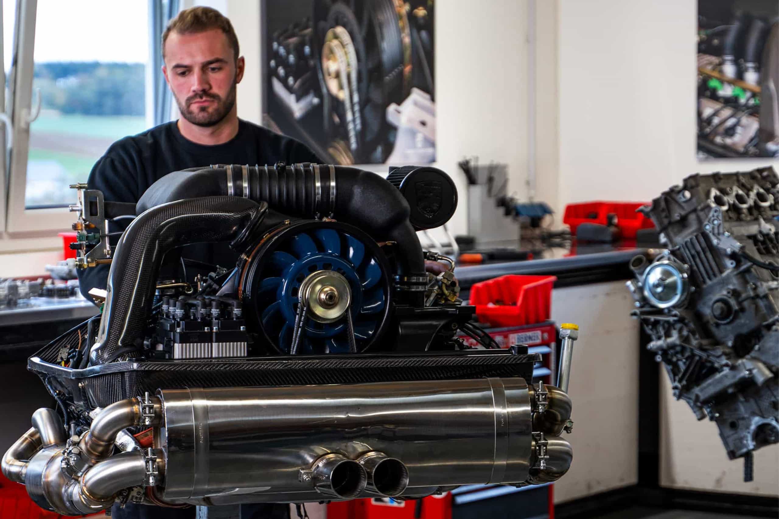 Ein detaillierter, auf einem Ständer montierter Automotor ist in einer Werkstatt ausgestellt, wobei zwei Personen im Hintergrund verschwommen sind. Der Raum hat graue Böden, weiße Wände und sichtbare Holzbalken an der Decke.