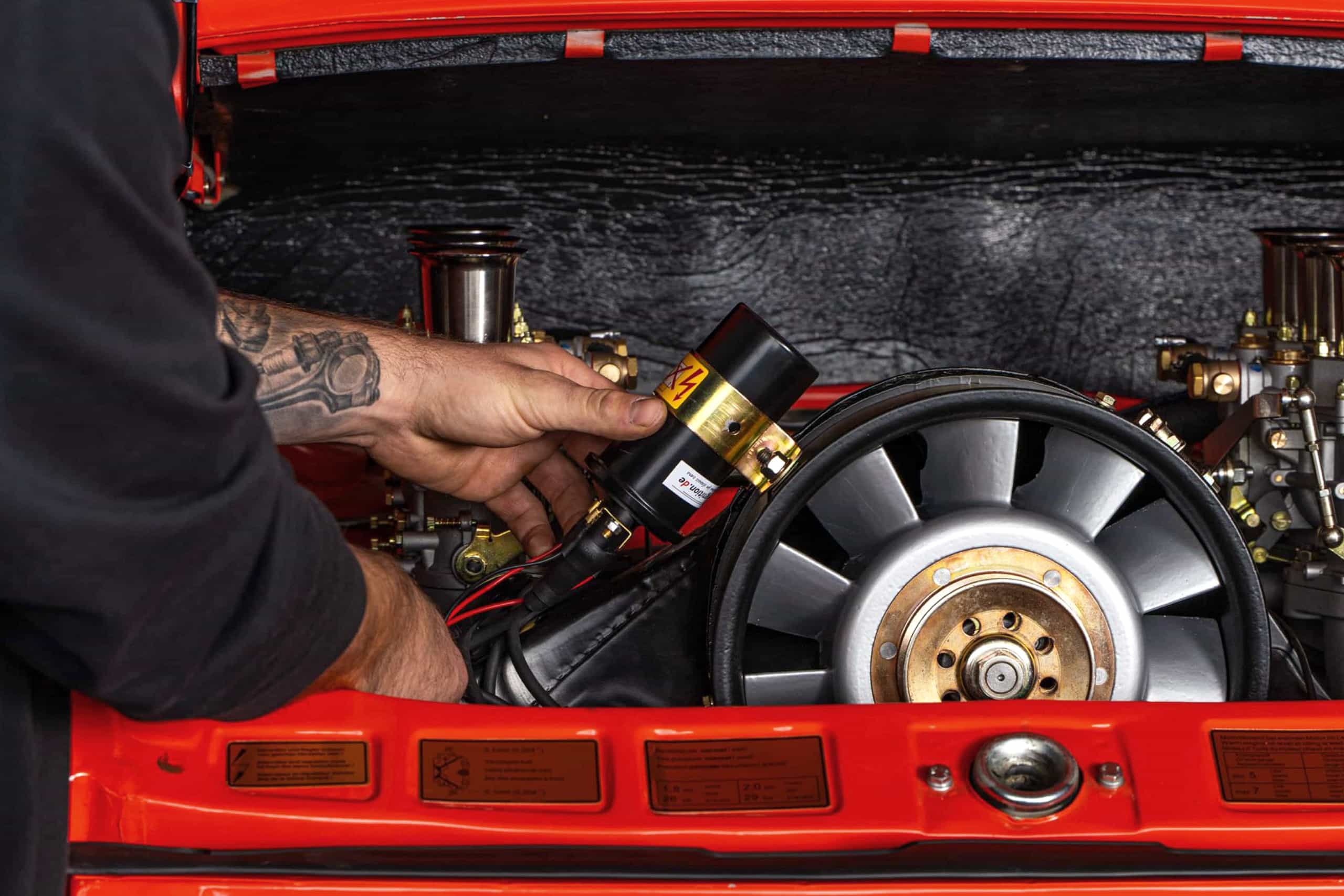 Eine Person mit tätowierten Armen arbeitet am freiliegenden Motor eines roten Oldtimers und stellt ein Bauteil im offenen Kofferraum ein oder baut es ein.