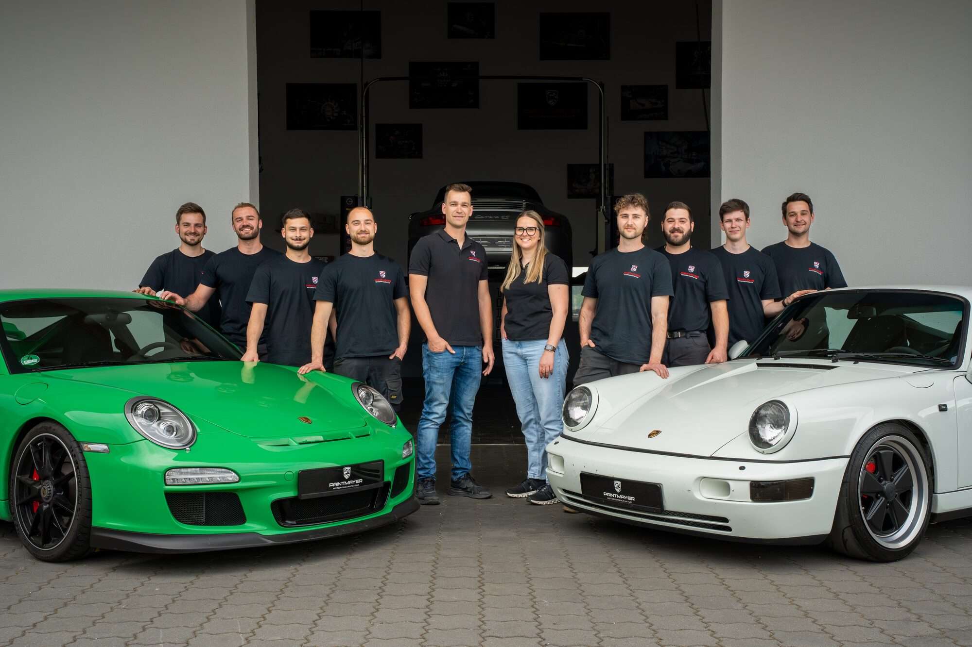 Drei Automechaniker arbeiten in einer hellen Garage an Luxusautos: Einer gießt Flüssigkeit in den Motor eines schwarzen Autos, ein anderer reinigt Motorteile, und ein dritter inspiziert die Unterseite eines gelifteten grünen Sportwagens.